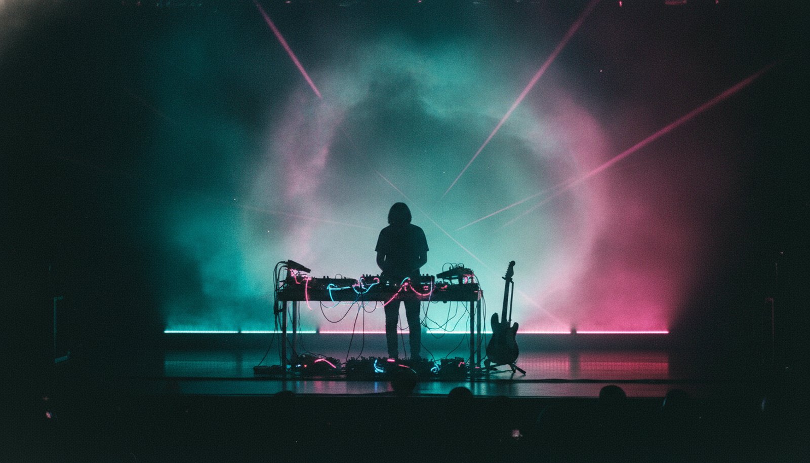 A solo live-electronic musician in silhouette behind glowing synths and a guitar, lit teal and magenta