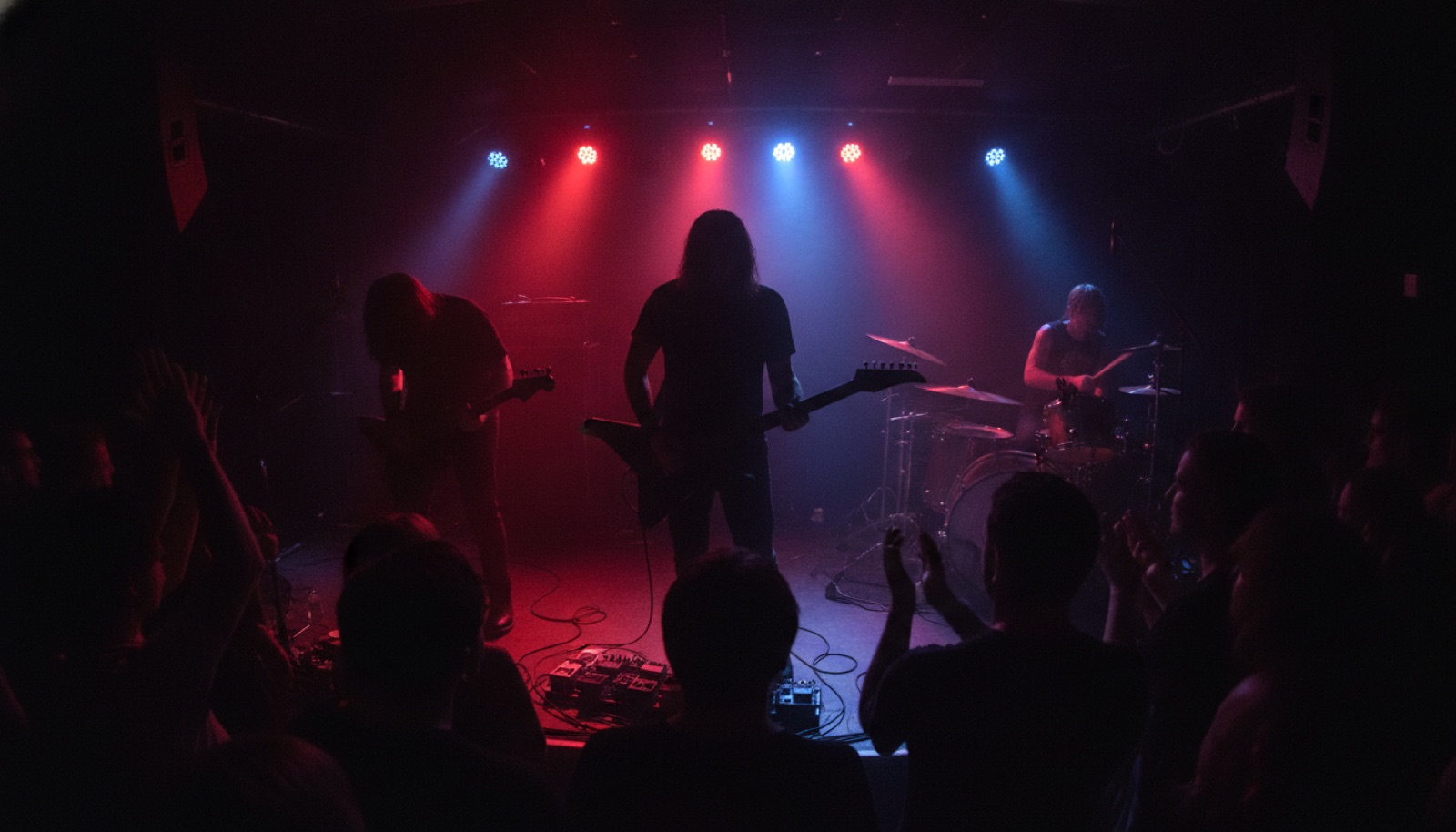 A cramped, sweat-lit rock club stage viewed from the crowd, guitars in silhouette under warm stage lights